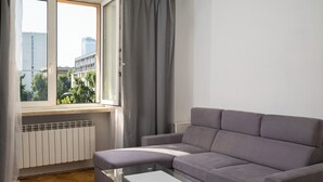 A modern living room with a grey sofa, a glass coffee table, and a view of buildings through a window with grey curtains.