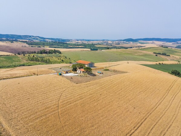 An aerial view of a rural landscape with rolling hills, fields, and a small building with a red roof surrounded by a fenced area.