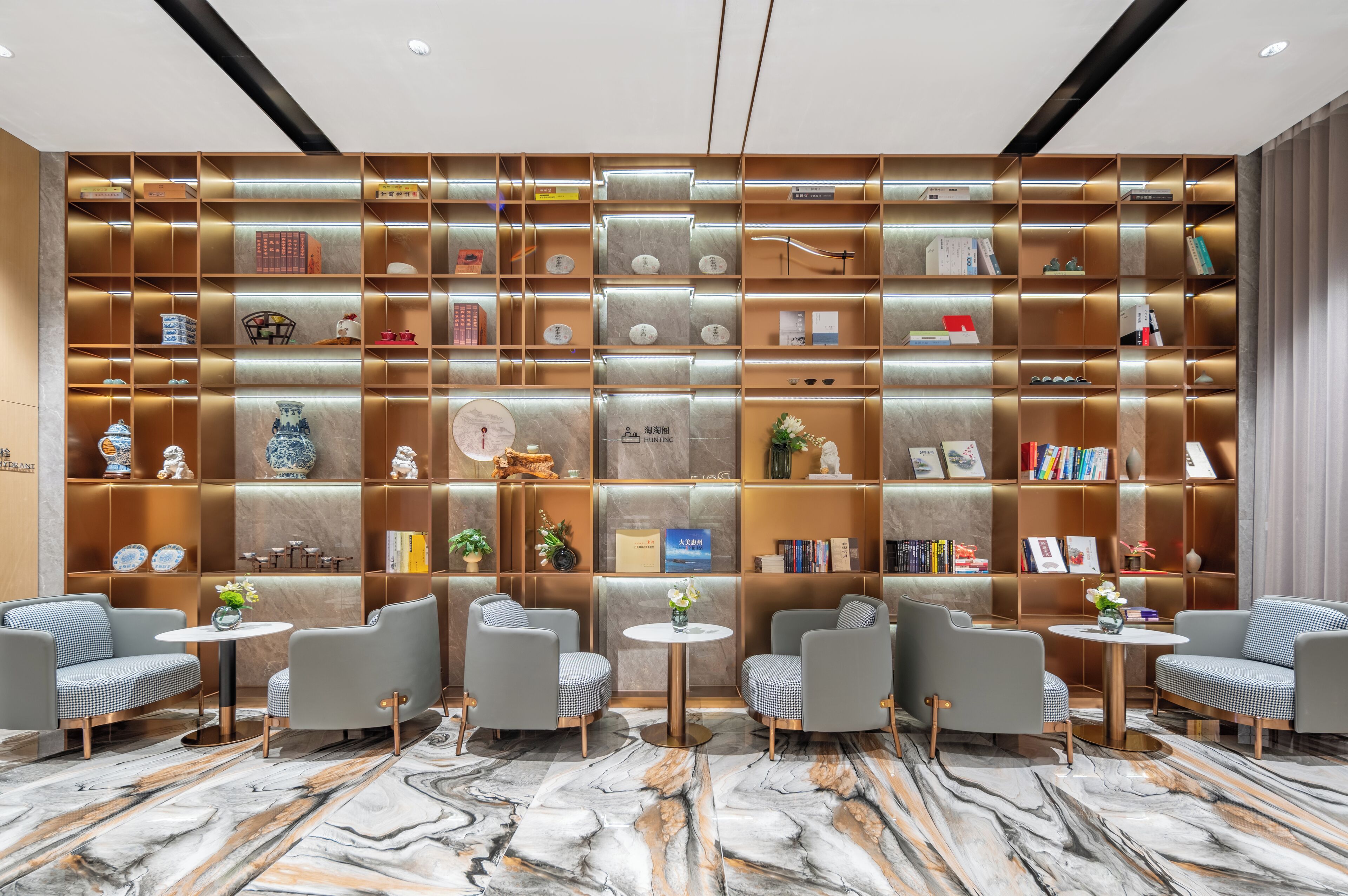 Un hall d’hôtel moderne avec un sol en marbre, une grande bibliothèque remplie de livres et d’objets décoratifs, et deux tables rondes avec des chaises.