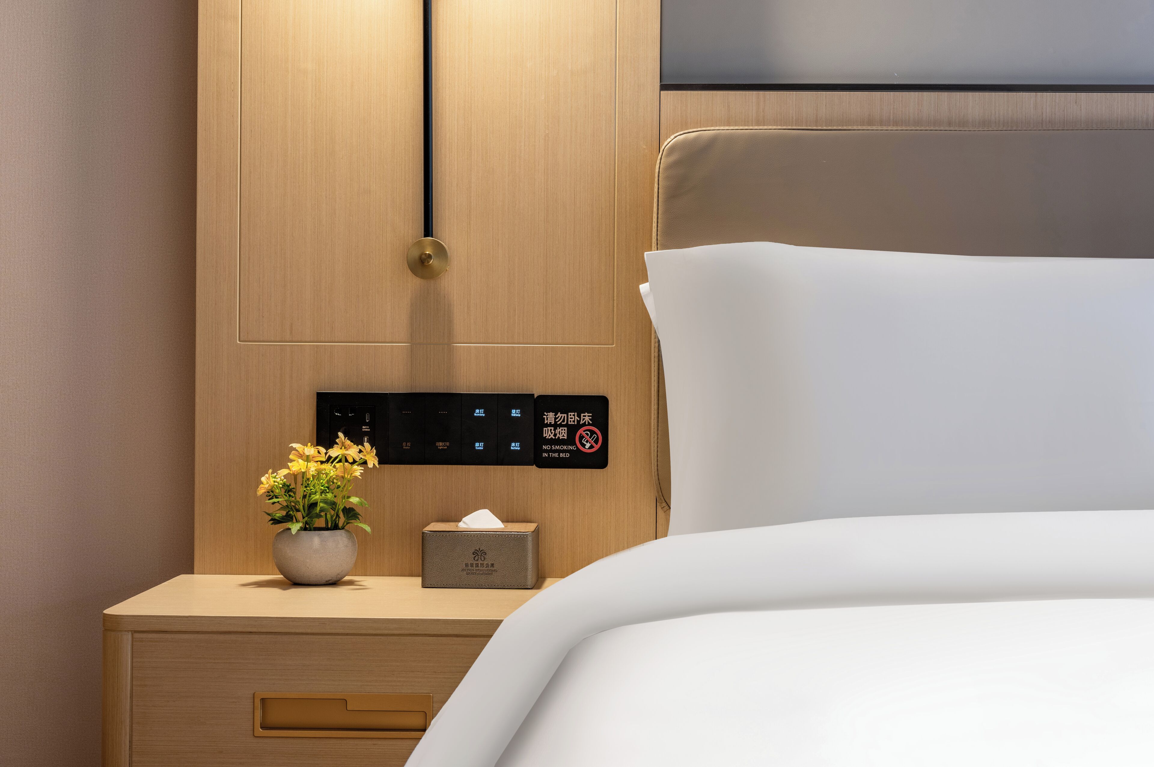 A hotel room with a wooden nightstand, a vase of flowers, a tissue box, and a digital control panel.
