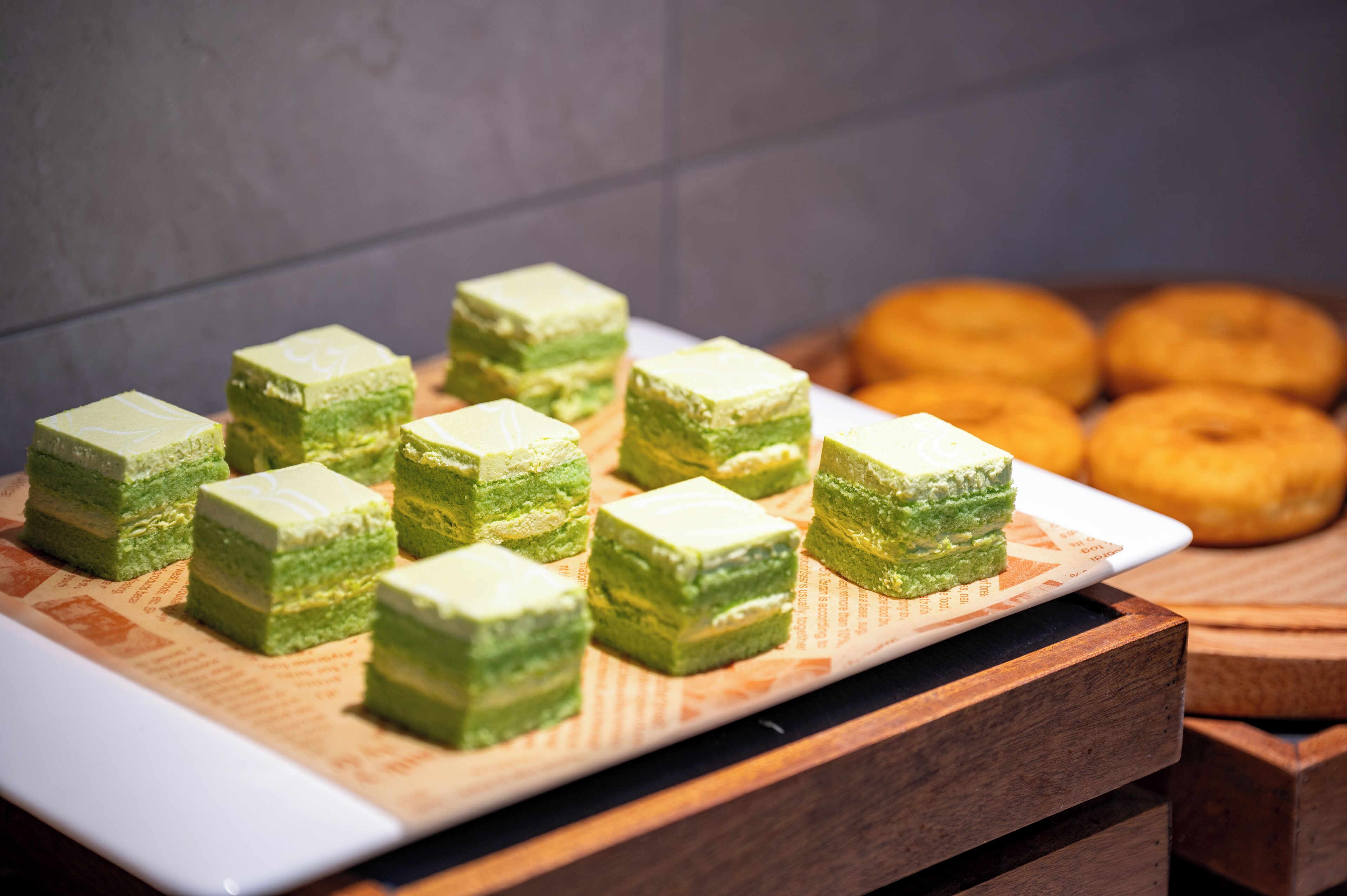 A tray of green layered cakes on a white plate, with a wooden table and a blurred background.