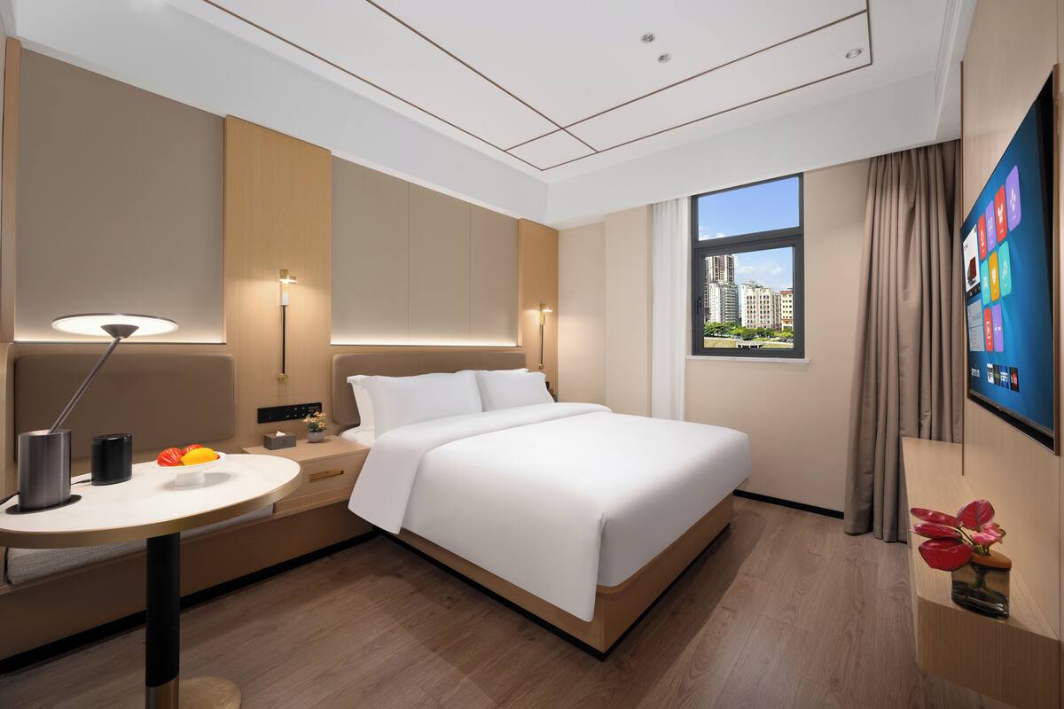 Habitación de hotel moderna con una cama grande, mesita de noche, lámpara y vistas a la ciudad a través de una ventana.