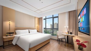 A modern hotel room with a large bed, a desk, a chair, a TV, and a window with a city view. - Platinum International Apartments (Huiyang High speed Railway Station Xinghe COCO Store) (Huizhou)