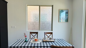 A dining area with a checkered tablecloth, two wooden chairs, a window with frosted glass, and a framed picture on the wall.
