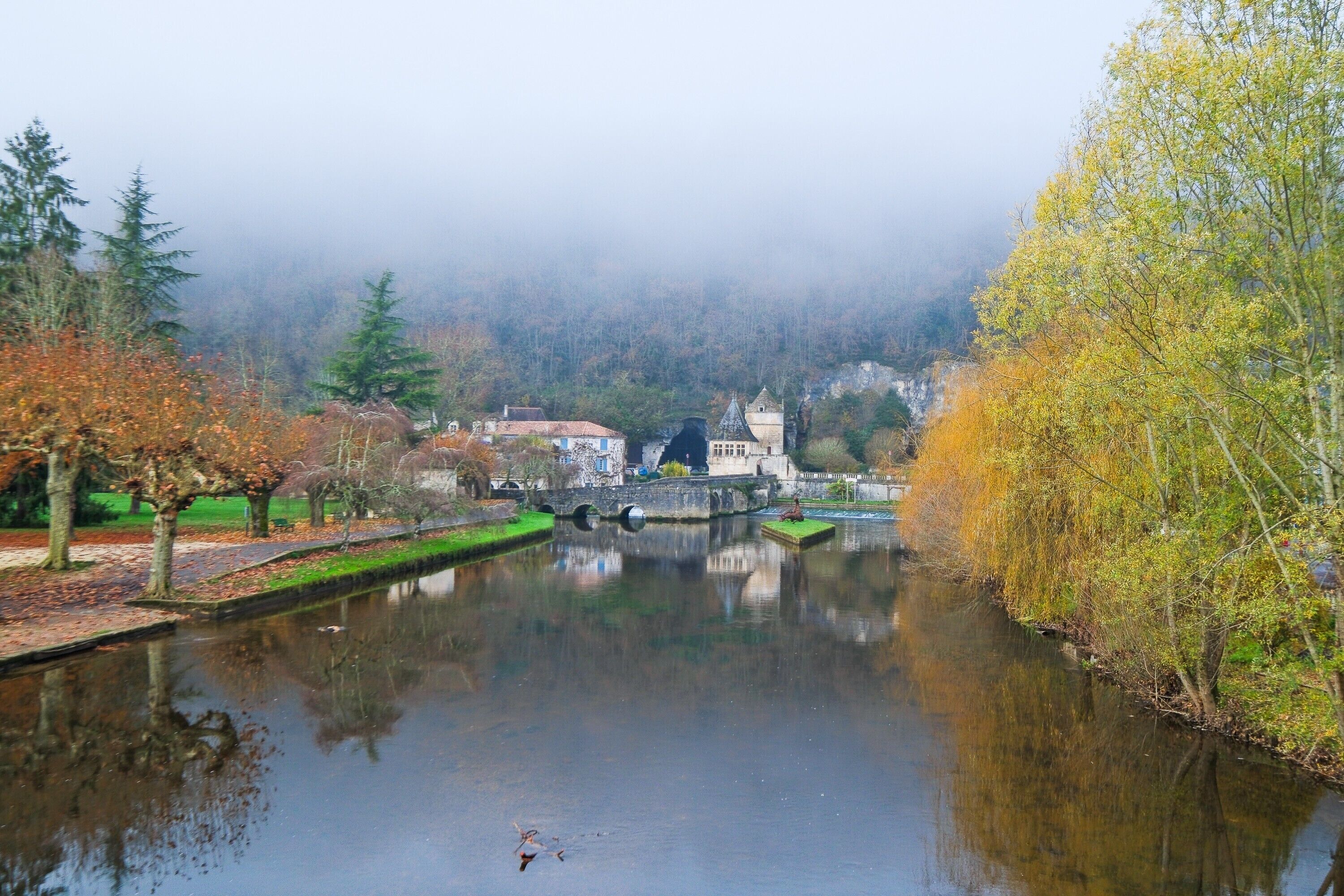 A calm river with a bridge, trees, and buildings in the background.