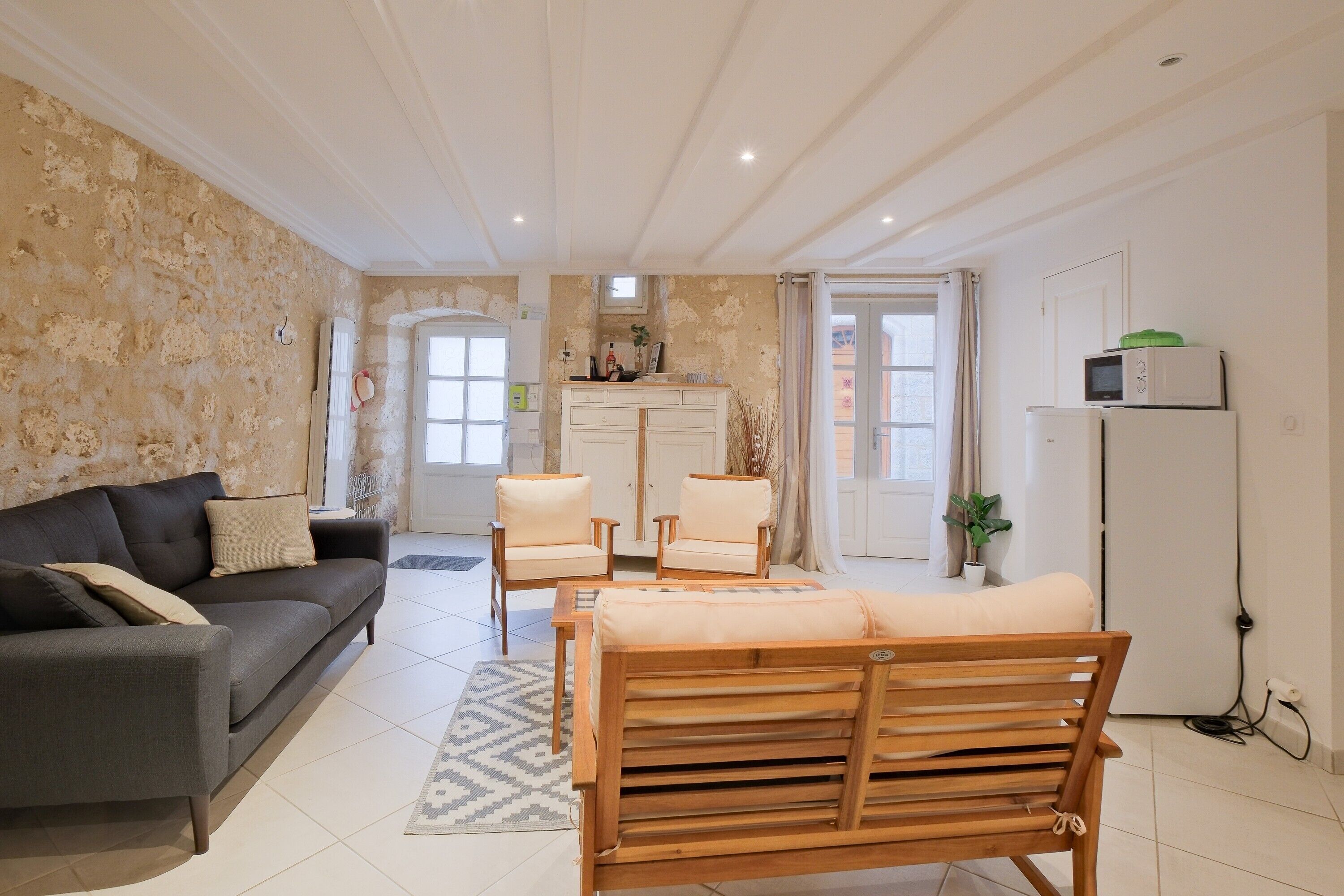 Un salon comprenant un canapé, deux fauteuils, un banc en bois, un coin cuisine avec un réfrigérateur et une porte donnant sur une autre pièce.