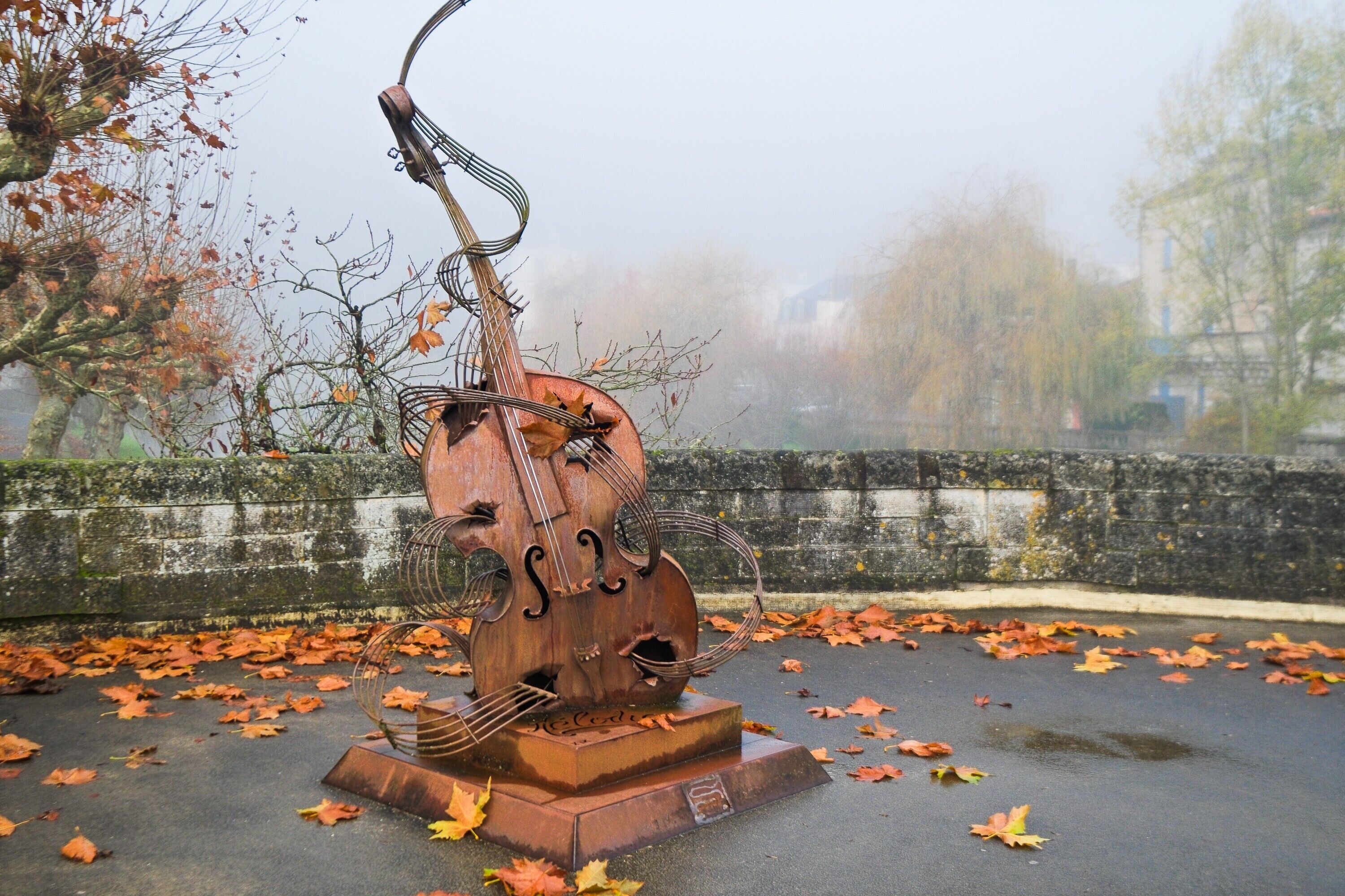 Een beeldhouwwerk van een cello op een voetstuk, omringd door gevallen bladeren.
