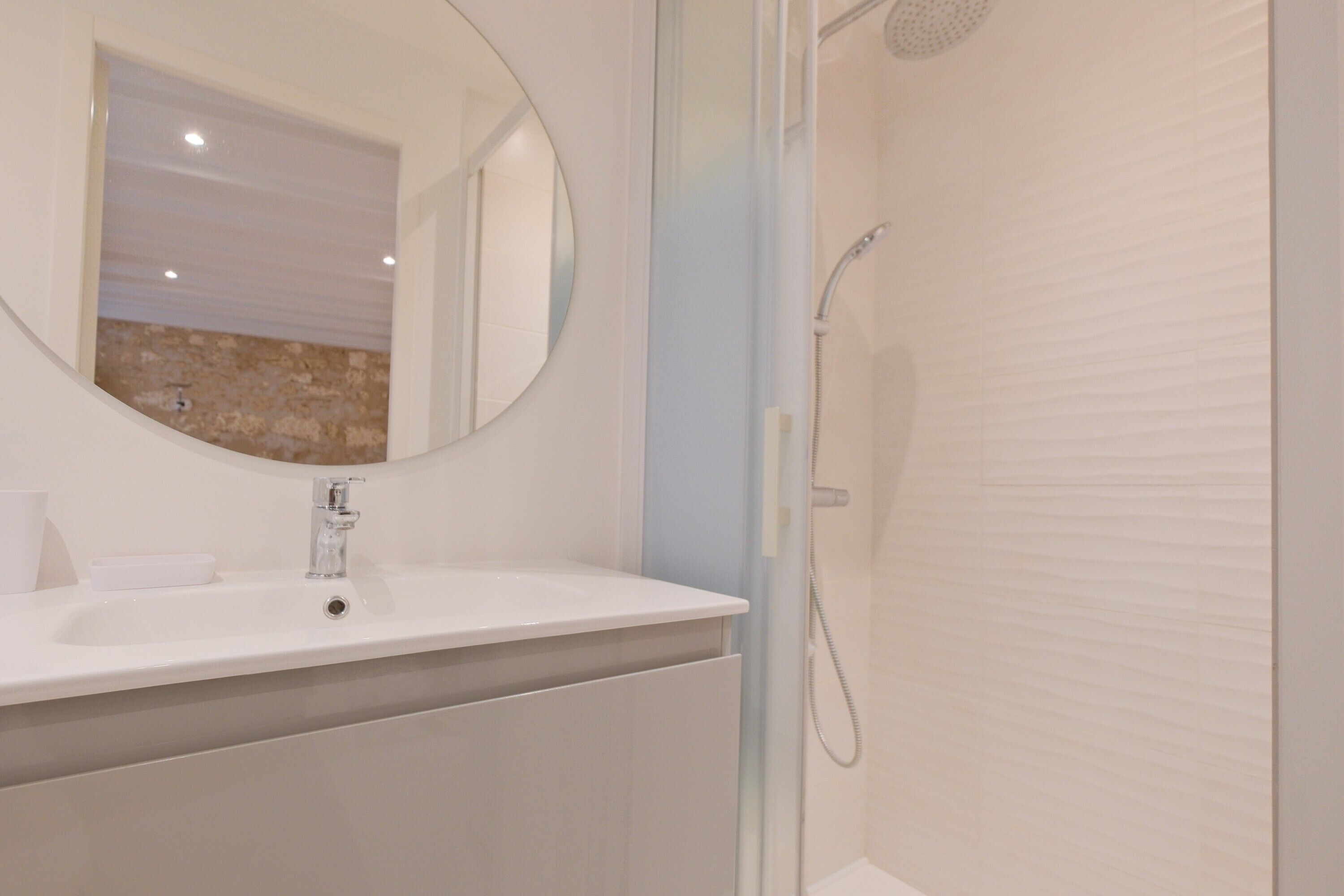 Une salle de bain avec un lavabo blanc, un miroir rond, un espace douche et un mur en pierre en arrière-plan.