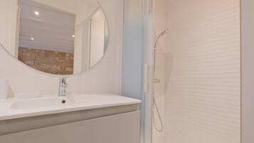Une salle de bain avec un lavabo blanc, un miroir rond, un espace douche et un mur en pierre en arrière-plan.
