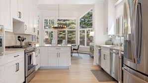 A modern kitchen with white cabinetry, stainless steel appliances, and large windows offering a view of the outdoors. - Luxury Home - Walk to Arrow Bahn Lift - 6-Bedrooms with 4 Kings & Game Room (Edwards)