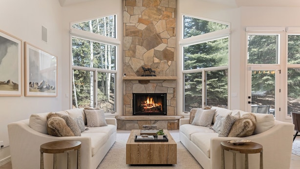 A modern living room with two white sofas, a stone fireplace, and large windows offering a forest view. - Luxury Home - Walk to Arrow Bahn Lift - 6-Bedrooms with 4 Kings & Game Room (Edwards)