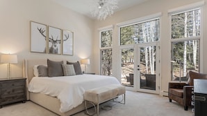 A spacious bedroom with a large bed, multiple pillows, a nightstand, and a desk with a chair. There are three large windows providing a view of the outdoors. - Luxury Home - Walk to Arrow Bahn Lift - 6-Bedrooms with 4 Kings & Game Room (Edwards)