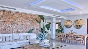 Una sala de estar moderna con una pared de piedra, una mesa de centro de vidrio, un sofá blanco y una zona de comedor con mobiliario de madera.