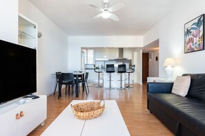 Un salon moderne avec un téléviseur à écran plat, une table basse, un panier en osier, un canapé en cuir noir, un coin repas avec des chaises et une cuisine en arrière-plan.