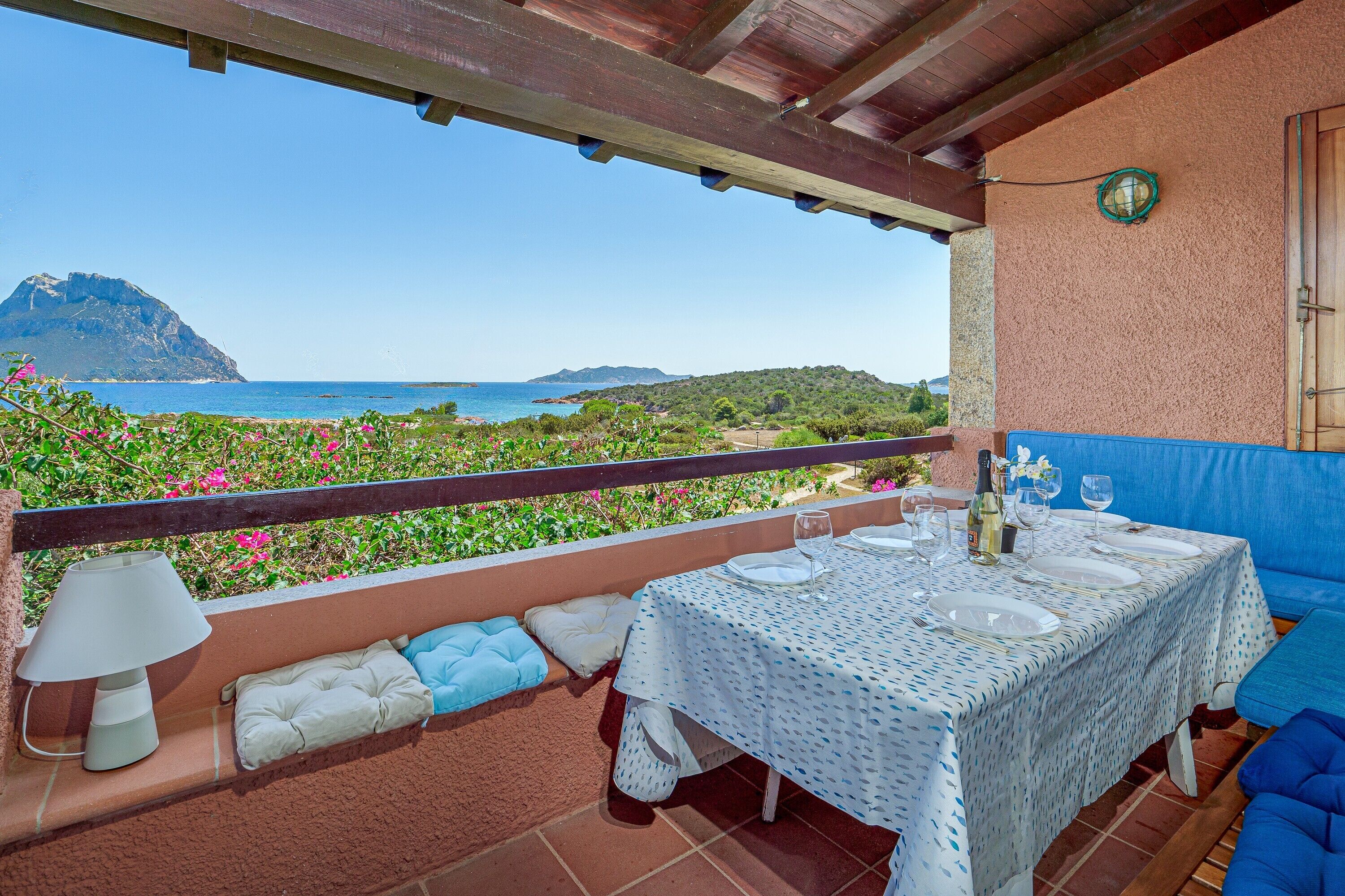 A terrace with a table set for dining, overlooking a coastal landscape with mountains and a clear sky.