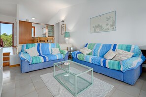 A living room with two blue sofas, a glass coffee table, a dining area with a wooden table and chairs, and a kitchen in the background. - Apartment 'Casa Sardina' with Sea View, Private Terrace (Loiri Porto San Paolo)