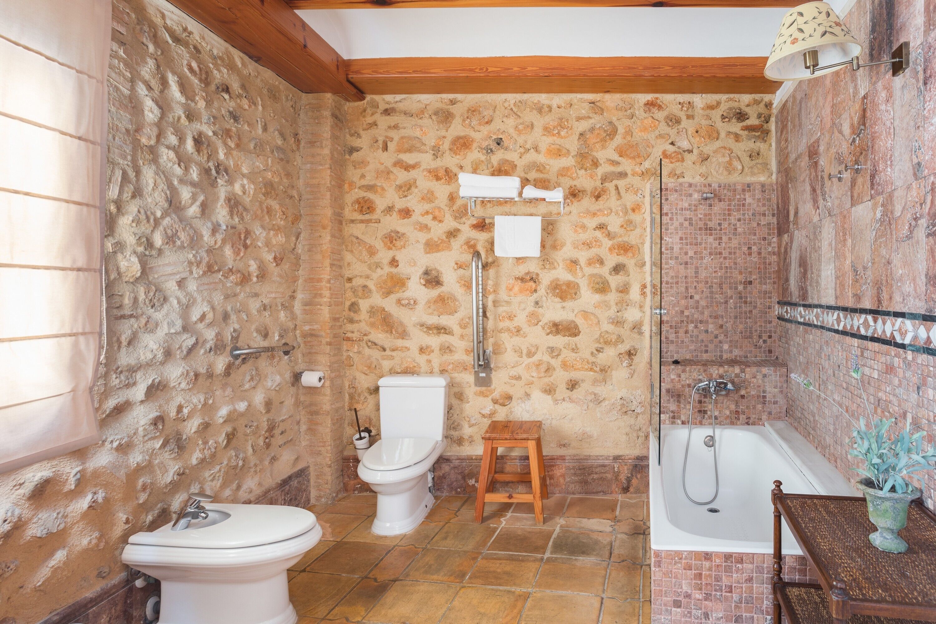 Een badkamer met een stenen muur, een toilet, een wastafel, een houten krukje, een bad en een handdoekhouder.