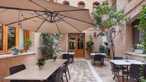 Zona de terrassa a l'aire lliure amb taules, cadires, un parasol gran i plantes en testos.