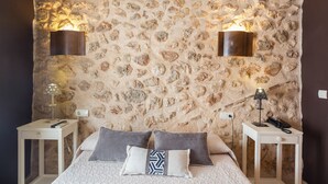 A bedroom with a stone wall, two bedside tables with lamps, a bed with pillows, and a nightstand with a phone. - Apartment 'Habitacion Estandar - Edificio Contiguo' with Private Terrace, Wi-Fi and Air Conditioning (Vilallonga)