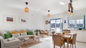 Una sala de estar moderna con sofá, mesa de comedor y sillas, una cocina con un ventanal grande, y un balcón con vistas.