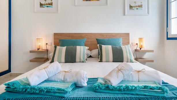 Una cama bien hecha con toallas y una manta azul, dos mesitas de noche con lámparas y cuadros en la pared.