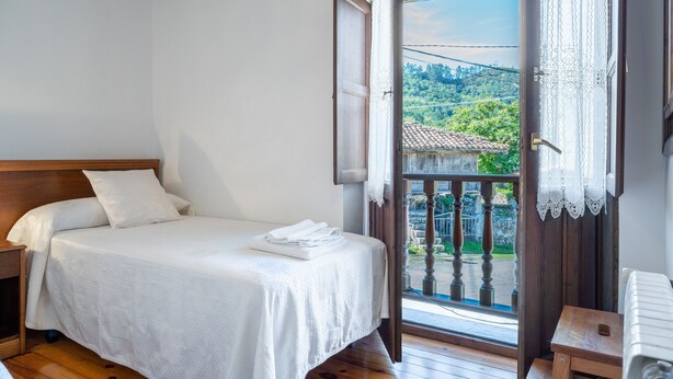 A bedroom with a bed, a nightstand, a wooden bench, a radiator, and a balcony with a view of greenery and a tiled roof.