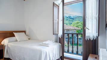 Een slaapkamer met een bed, nachtkastje, houten bankje, radiator en balkon met uitzicht op groen en een pannendak.