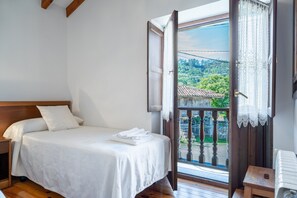 A bedroom with a bed, a nightstand, a wooden bench, a radiator, and a balcony with a view of greenery and a tiled roof.
