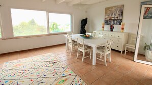 A dining area with a white table, chairs, a rug, and a window with a view of greenery. - Holiday Home 'Villa Betty' with Sea View, Wi-Fi and Air Conditioning (Dénia)