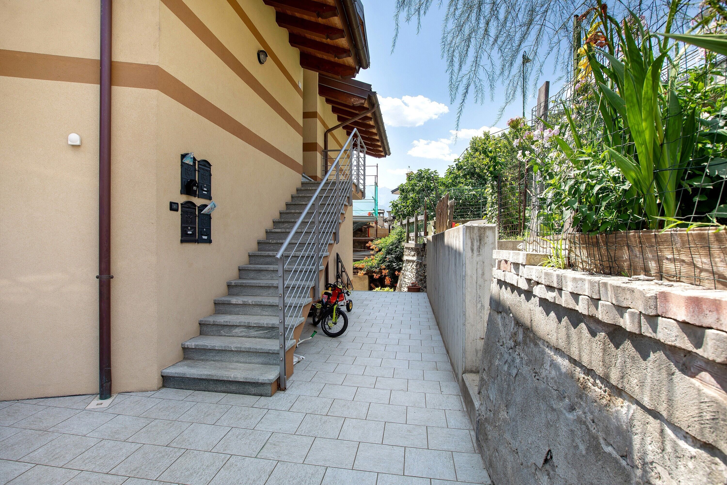 Un étroit couloir extérieur avec un escalier métallique menant à l’entrée d’un bâtiment, un vélo garé et un mur dont la base est en briques.