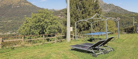 Een gazon met een metalen prieel, een tafel met een blauwe hoes en twee ligbedden.