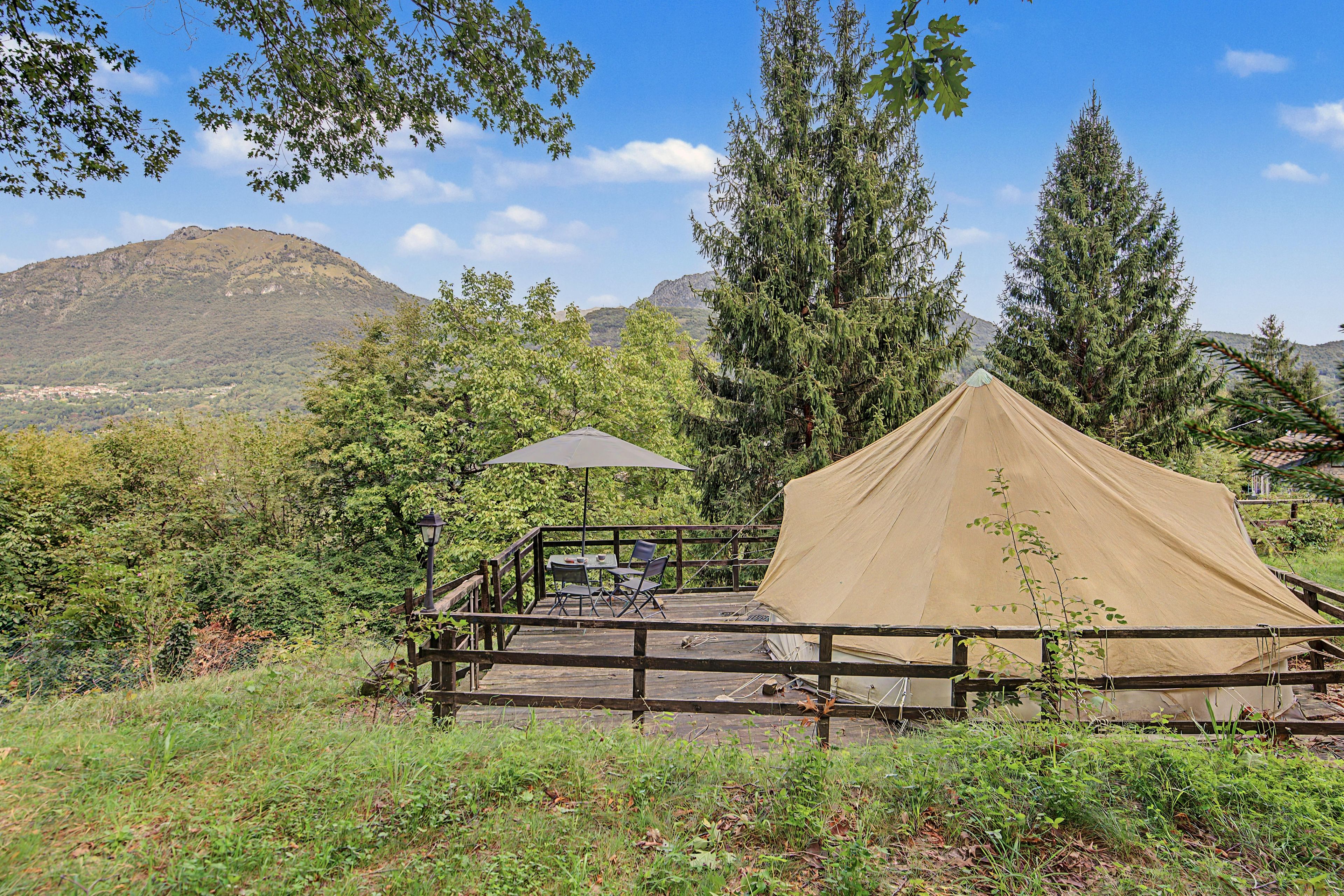 Un espacio para acampar con una tienda de campaña, una mesa y sillas, rodeado de árboles y una montaña al fondo.