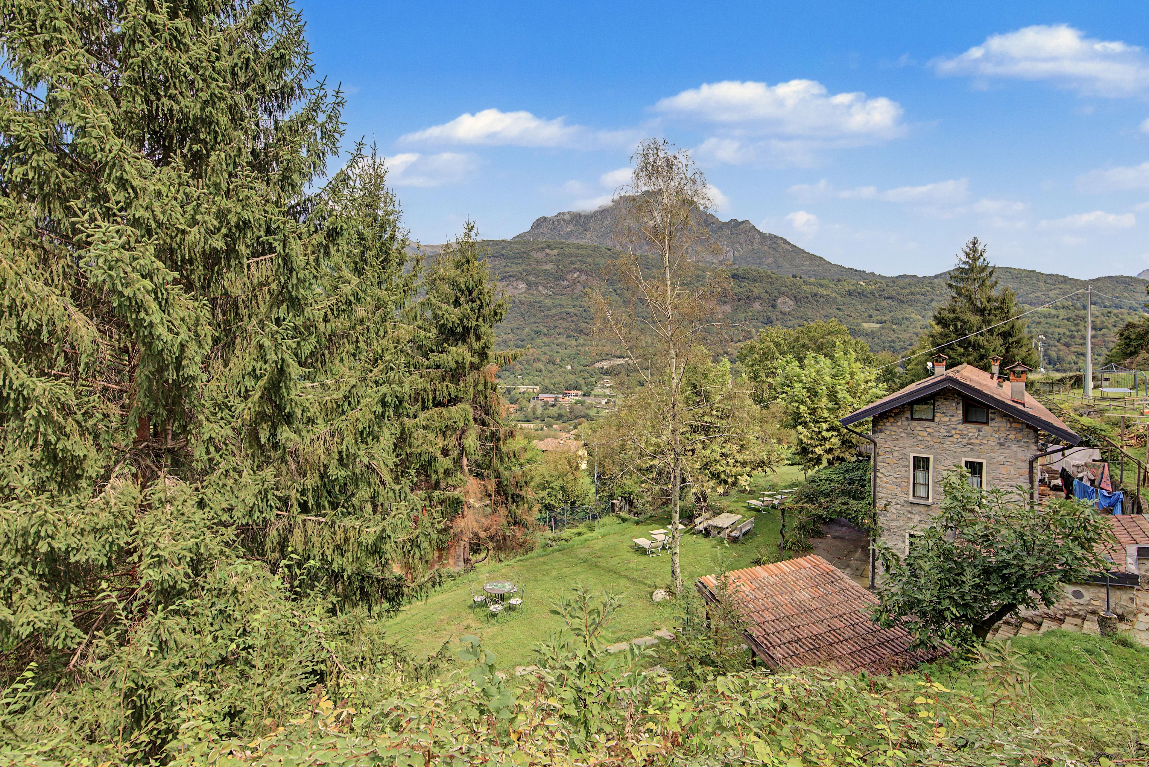 Paesaggio rurale con una casa in pietra, un campo erboso e una montagna sullo sfondo.