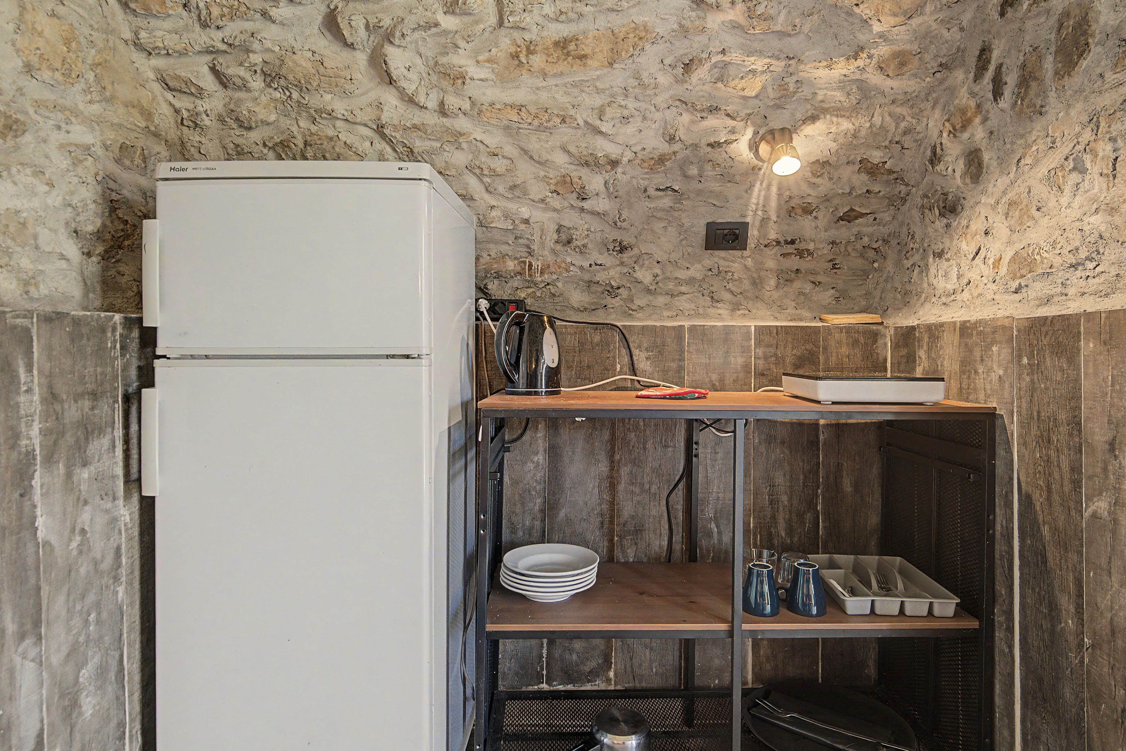 A compact kitchen with a white refrigerator, a wooden shelf, and a metal rack with dishes.