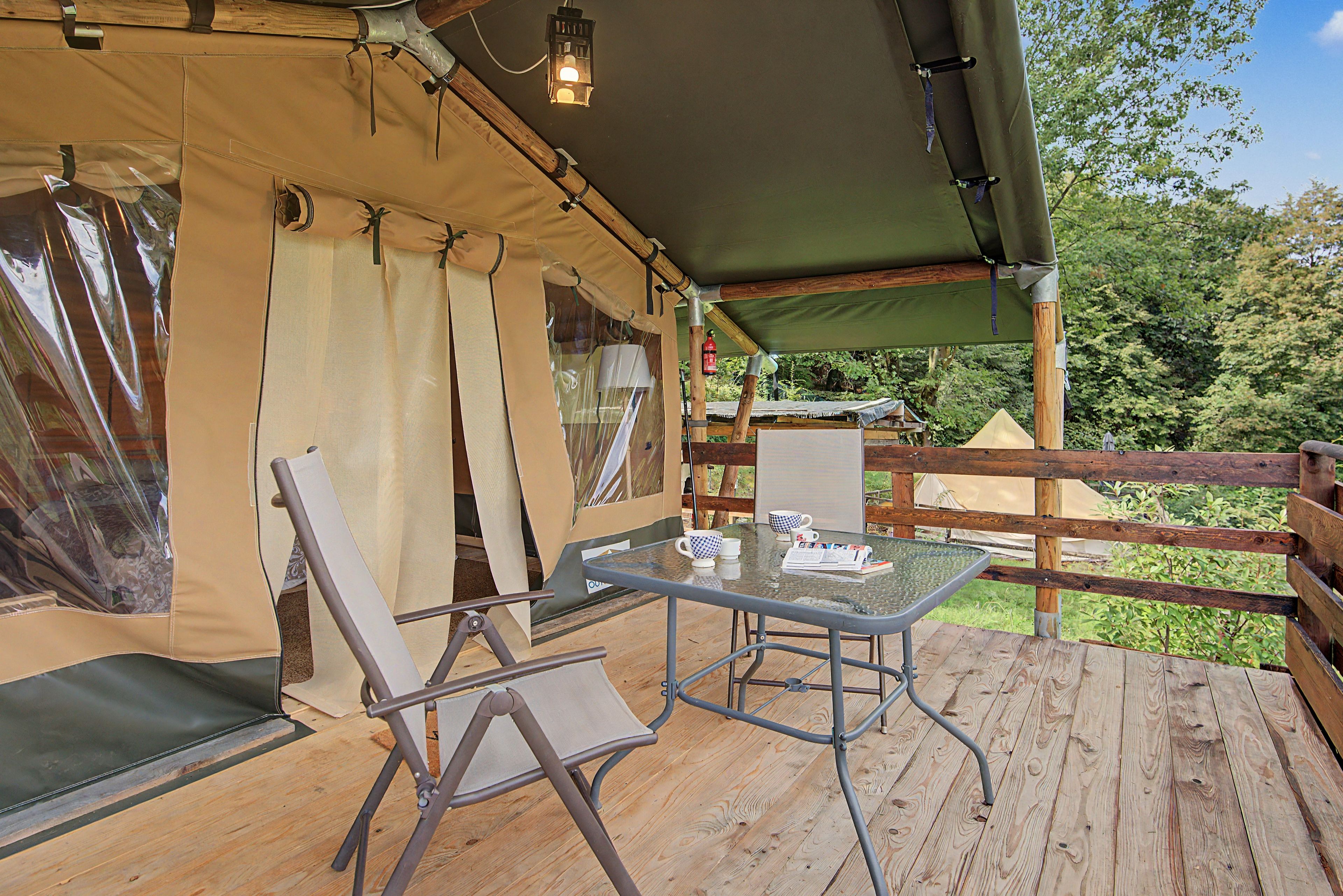 Eine Holzterrasse mit einem Glastisch und Stühlen, eine mit Vorhängen versehene Konstruktion und Blick auf Bäume.