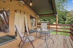 Eine Holzterrasse mit einem Glastisch und Stühlen, eine mit Vorhängen versehene Konstruktion und Blick auf Bäume.