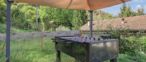 Una mesa de fútbol de mesa al aire libre bajo una cubierta, rodeada de follaje y una valla de madera.