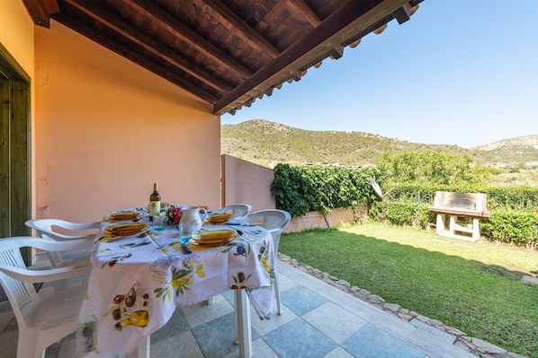Une terrasse extérieure avec une table dressée pour le repas, entourée de chaises blanches, d’une pergola en bois et offrant une vue sur un paysage vallonné.