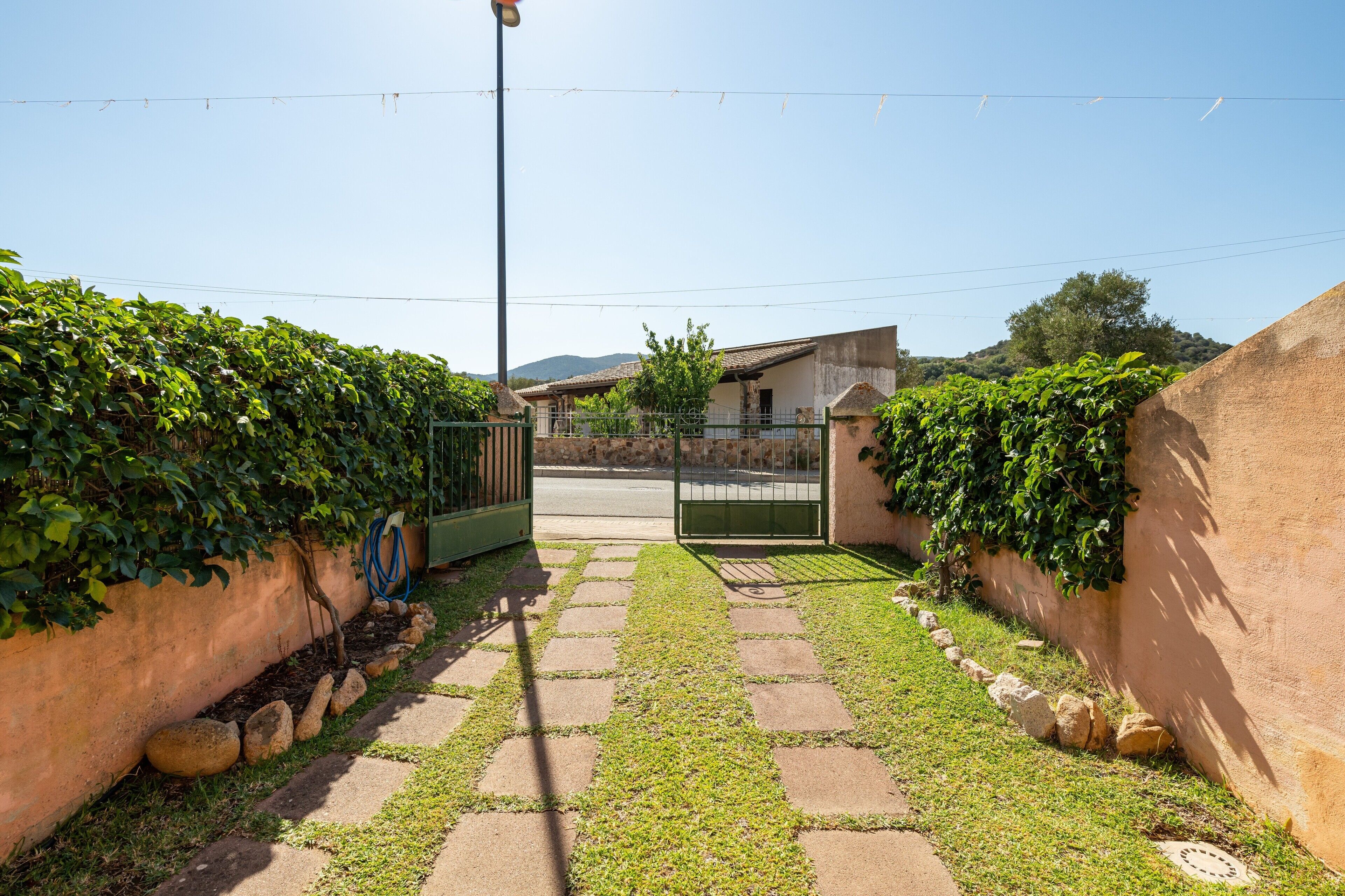 A paved pathway leading to a gated entrance, flanked by walls with climbing plants, and a clear sky above.