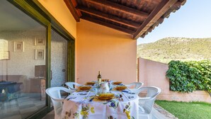 Un área de comedor al aire libre con una mesa preparada para cuatro personas, rodeada de sillas blancas, un ventanal de vidrio que da al interior y vistas a una colina.