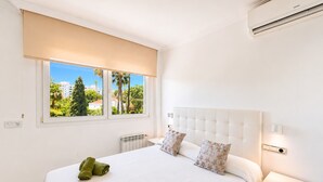 A hotel room with a bed, two windows, a radiator, a nightstand, and an air conditioning unit. - Villa 'Gran Cerca De Puerto Banús' with Mountain Views, Wi-Fi, and Air Conditioning (Marbella)