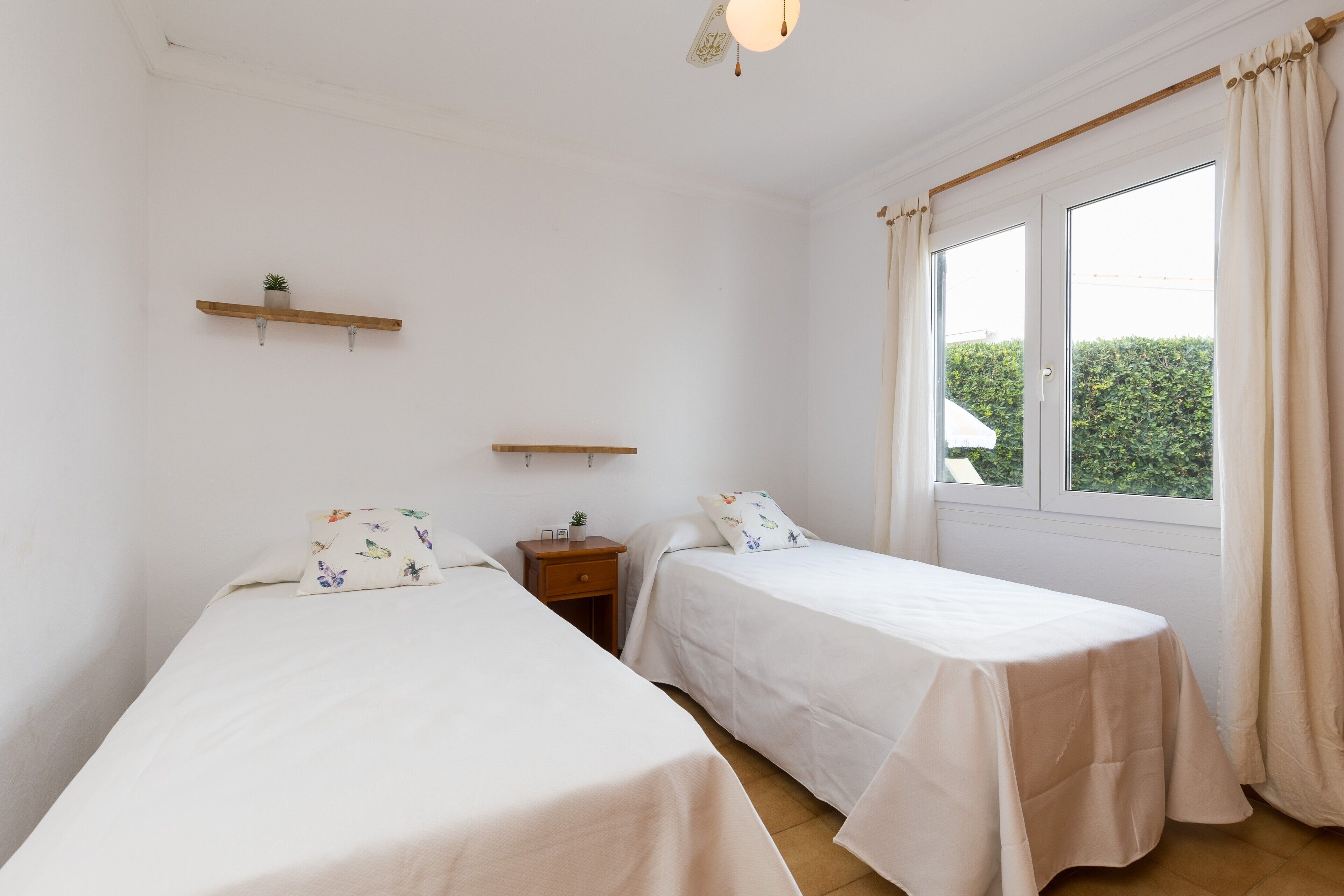 Une chambre avec deux lits, une petite table, une table de chevet, une étagère avec une plante et une fenêtre avec des rideaux.