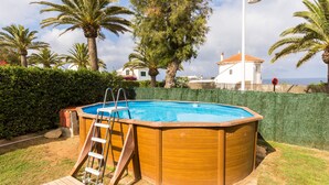 Uma banheira de hidromassagem de madeira com uma escada, cercada por um sebedeiro e palmeiras, com uma casa e o oceano ao fundo.