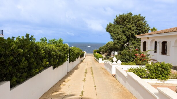 Rua estreita que leva a um corpo d'água com um barco, cercada por edifícios brancos e vegetação.