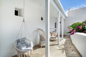 Un espace extérieur aux murs blancs, agrémenté d’un fauteuil suspendu, de fauteuils en osier et d’un petit jardin où poussent des fleurs roses.