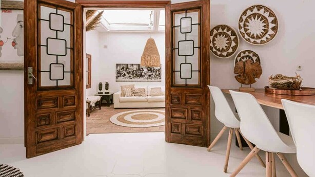 Una habitación con puerta de madera, una mesa de comedor con sillas, un sofá, un tapete estampado y adornos en la pared.