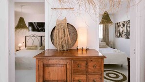 Un dormitorio con un aparador de madera, un espejo redondo, una decoración colgante de tejido, una cama con colcha blanca y una obra de arte colgada en la pared.