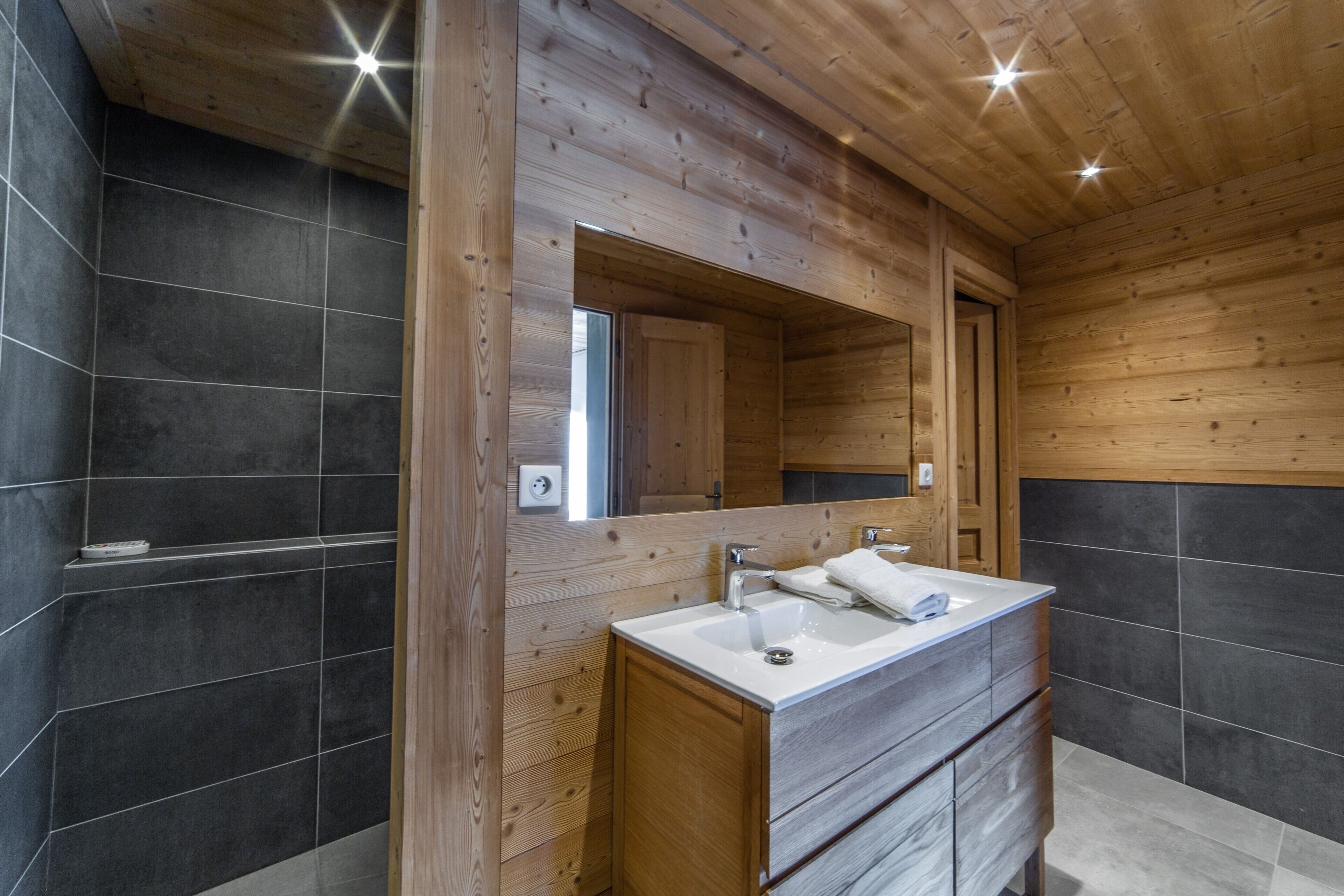 A modern bathroom with a wooden ceiling, a large mirror, a double sink, and dark tiled walls.