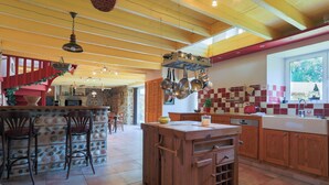 A kitchen with wooden cabinets, a central island, hanging pots, and a red staircase. - Holiday Home 3 min from Beach – Private Terrace, Fenced Garden, Sleeps 5 (Plouha)