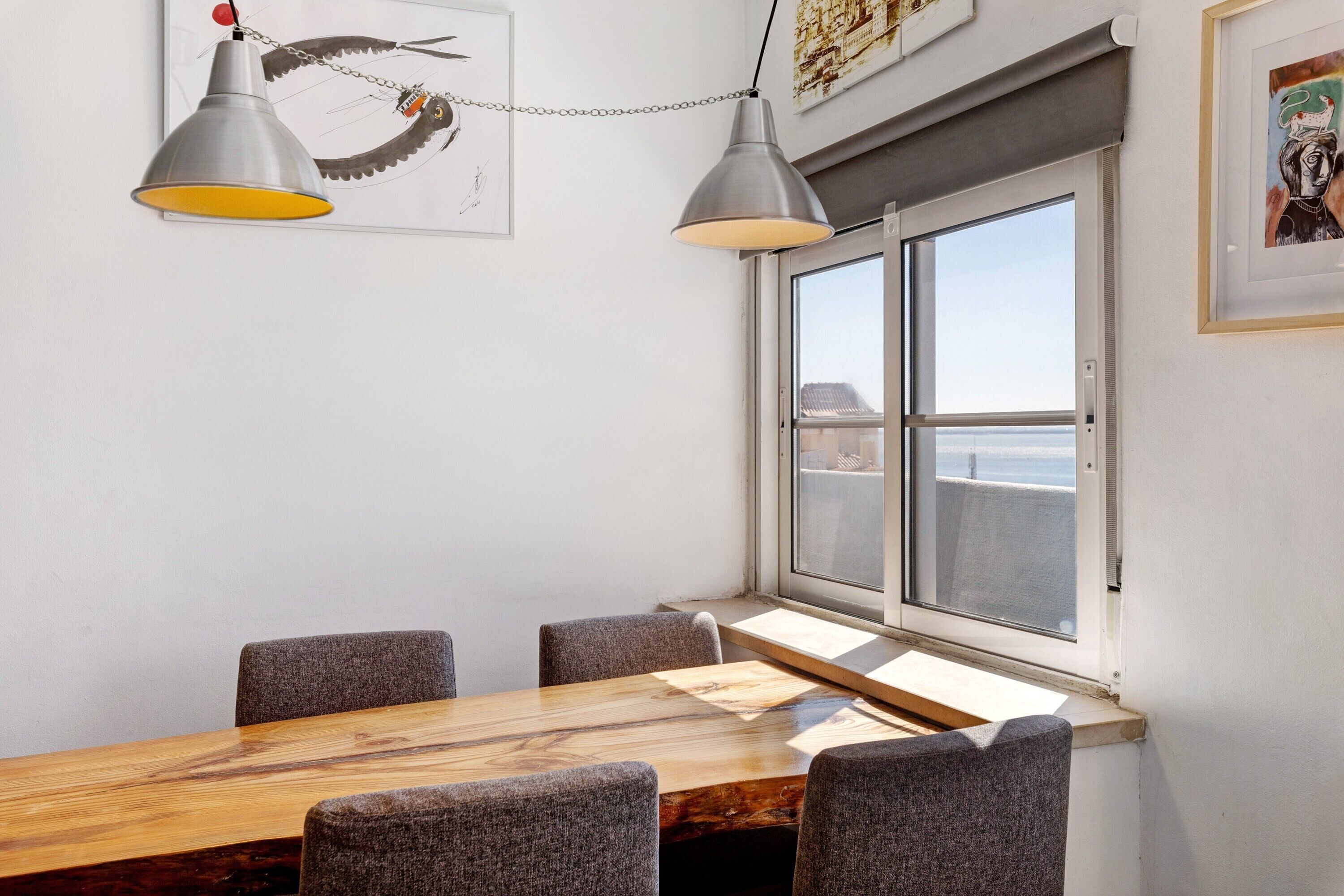 Ein Essbereich mit einem Holztisch, vier Stühlen, zwei Pendelleuchten, einem Fenster mit Ausblick und Bildern an den Wänden.
